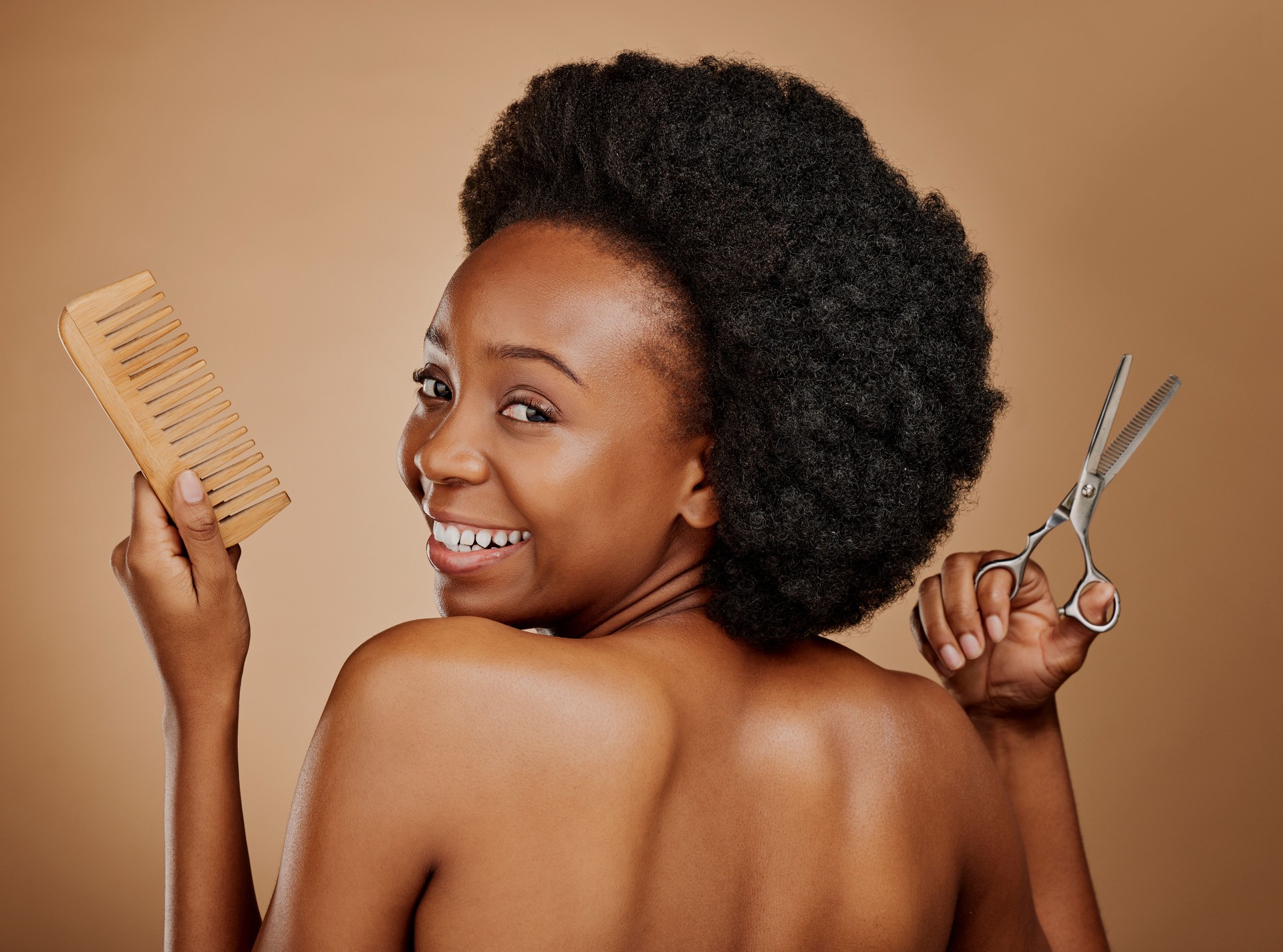 Portrait, back and a black woman with a comb and scissors in studio on a brown background for a haircut. Smile, afro and haircare with a happy young female person looking excited at the salon